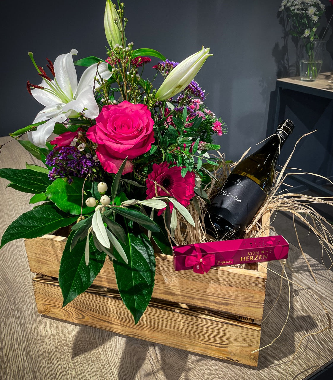 Blumenstrauß mit rosa Blüten zum Valentinstag, üppig gebunden, dazu eine Flasche Prosecco, eine kleine Pralinenbox. Alles arrangiert in einer geflammten Holzkiste.